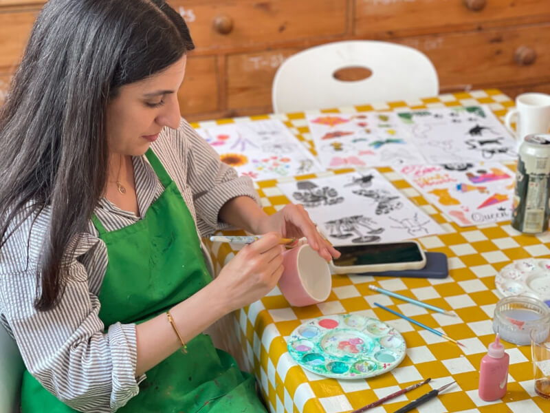 A woman is sitting at a table, painting a handmade ceramic cup pink